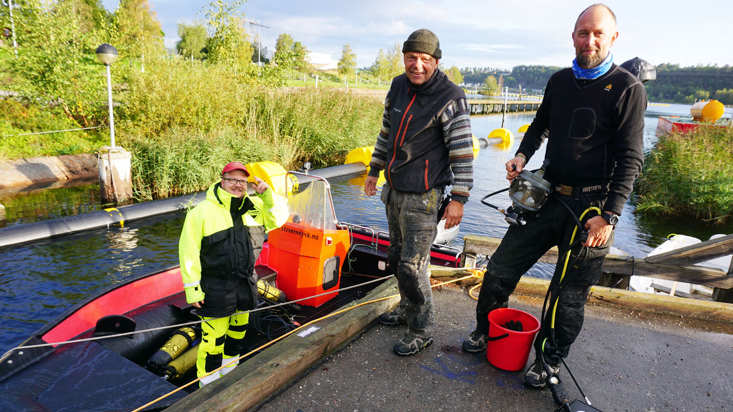 En mann i båt ved kai, to menn på kai, en av dem i dykkerdrakt.
