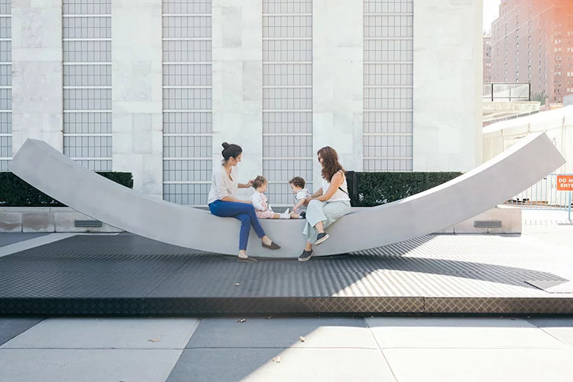 On the gently inwards sloping aluminium peace bench, two young mothers hold toddlers who play together.