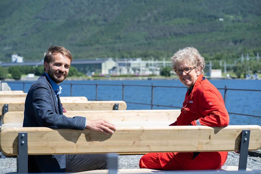 Vestre CEO Jan Christian Vestre and Hydro's CEO Hilde Merete Aasheim at a bench made using Hydro aluminium. Hydro's Husnes plant in the background. 