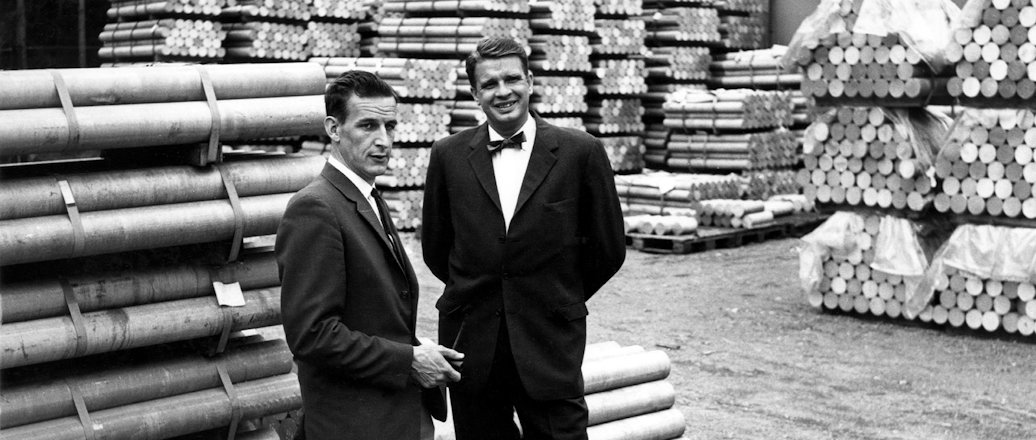 a couple of men standing in front of stacks of wood